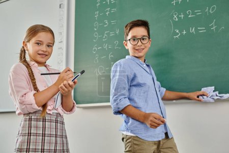 Foto de Niño y niña de pie frente a una pizarra, ansiosos por aprender en un salón de clases luminoso y animado. - Imagen libre de derechos