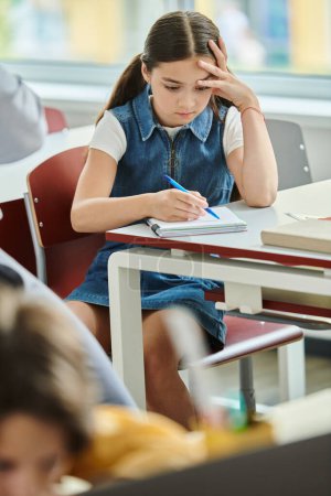 A young girl sits at a table with a notebook and pen, fully engrossed in her writing.