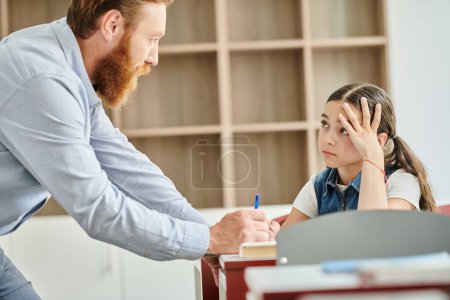 un enseignant, guidant une petite fille assise à un bureau dans un cadre de classe coloré