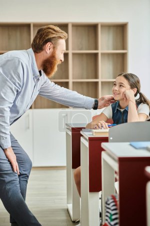 A red-bearded man shakes hands with a kid in a vibrant classroom full of diverse students, showcasing a warm and welcoming environment.