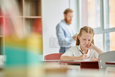 A young girl sits at a desk, engaged with a study in a bright, lively classroom while the teacher instructs behind her