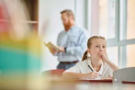 A young girl sits at a table while a man teacher stands behind her, instructing class