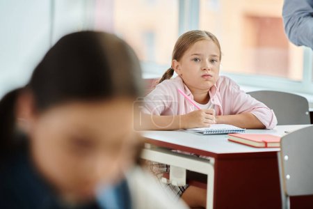 Une jeune fille avec des nattes s'assoit à son bureau, écoutant les instructions des professeurs dans une salle de classe animée.