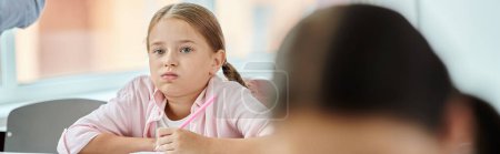 A young girl sits at a table, engrossed in her task, with a bored face expression in classroom setting.