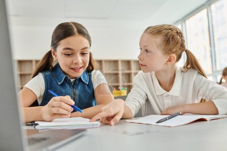 Photo for Two young girls with focused expressions sitting at a desk, diligently writing in a bright and lively classroom environment - Royalty Free Image