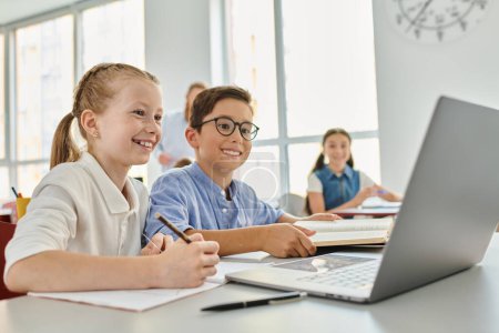 Téléchargez les photos : Un groupe d'enfants assis à une table, concentré sur un ordinateur portable dans une salle de classe lumineuse. - en image libre de droit