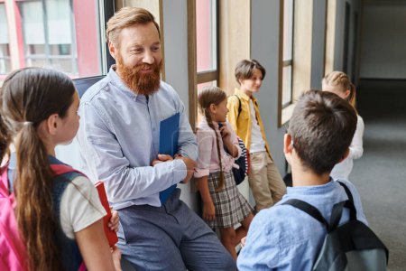 A bearded man stands confidently in front of a group of children in a vibrant classroom, engaging them in an educational activity.