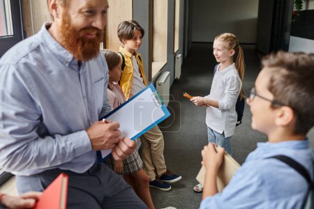 Foto de Un maestro varón sosteniendo un portapapeles está al lado de un grupo de niños en un ambiente vibrante en el aula. - Imagen libre de derechos