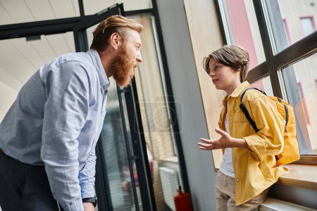A man stands beside a little boy in front of a window, engaging in a thoughtful conversation while looking outside.