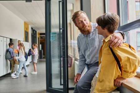 Foto de Un hombre con una ardiente barba roja mira atentamente a un joven vestido con una chaqueta de color amarillo brillante, posiblemente enseñándole o enseñándole. - Imagen libre de derechos