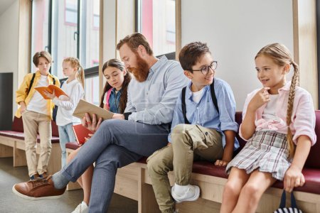 Téléchargez les photos : Groupe d'enfants et d'enseignants adultes assis, bavardant et écoutant attentivement sur un banc dans un cadre de classe animé et coloré. - en image libre de droit