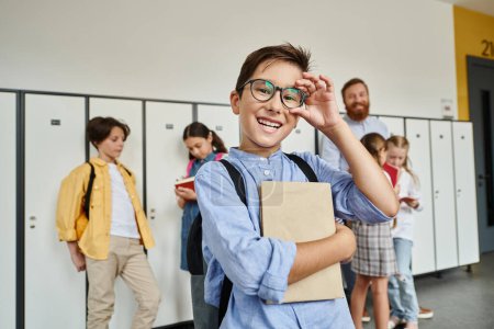 Un niño con una camisa azul y gafas se para con confianza frente a los casilleros en un pasillo de la escuela.