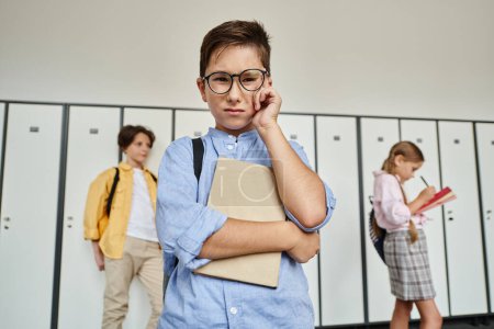 Un garçon en chemise bleue se tient au milieu des rangées de casiers dans un couloir de l'école.