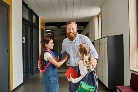 A bearded man is conversing with two attentive little girls in a vibrant classroom setting filled with energy and curiosity.