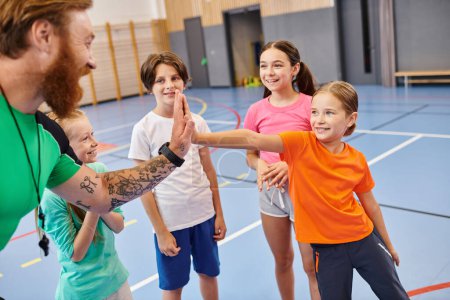 Un grupo diverso de niños pequeños se paran entusiasmados uno alrededor del otro en un entorno de gimnasio mientras escuchan las instrucciones de sus maestros varones..