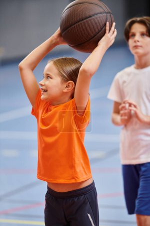 Téléchargez les photos : Une jeune fille tient joyeusement un ballon de basket haut dans les airs, irradiant un sentiment d'excitation et de passion pour le jeu. - en image libre de droit