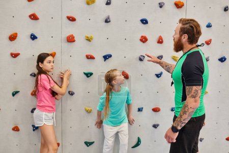 A male teacher is instructing two young girls standing in front of a climbing wall in a bright, lively classroom setting.