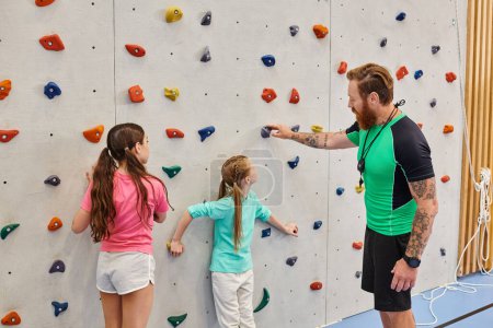 Un homme enseignant instruit deux jeunes filles devant un mur d'escalade dans un cadre de classe lumineux et animé.