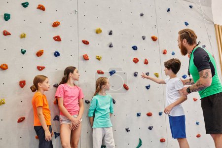 A man teaches a group of diverse children how to climb on a rock wall in a bright, lively classroom setting.