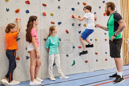 A diverse group of kids are energetically climbing and exploring a wall while being supervised by their male teacher in a bright, lively classroom setting.