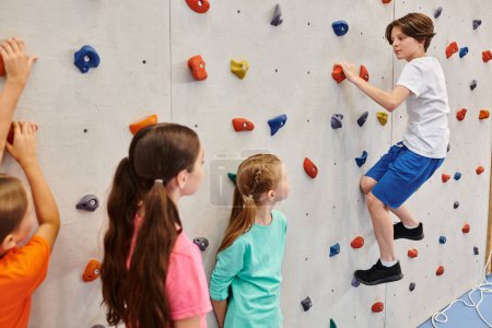 A group of young children stand together in front of a climbing wall, listening attentively to their teachers instructions before attempting to scale the wall.