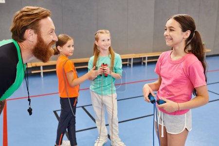 Un hombre barbudo enseña con entusiasmo a un grupo de niños diversos en un gimnasio vibrante, cautivando su atención con lecciones atractivas.