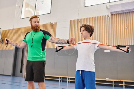 A bearded instructor with tattoos guides a young student through arm stretches in a vibrant school gymnasium.