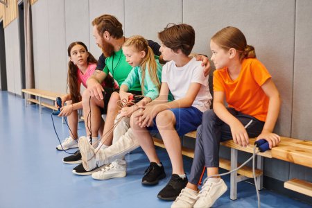 A diverse group of young children, seated on a bench, listening attentively as their male teacher imparts knowledge in a vibrant classroom setting.