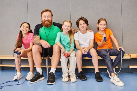 A man with a beard, a teacher, sitting on a bench surrounded by happy, diverse children of various ages, engaging in conversation and learning together.