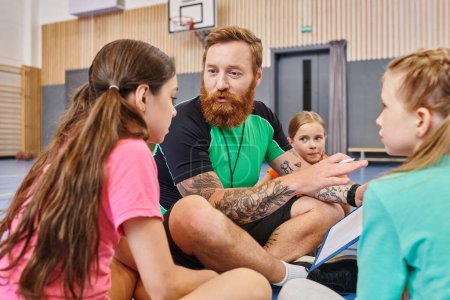 A bearded man sits on a basketball court surrounded by diverse children, engaging them in an inspiring lesson about the game.