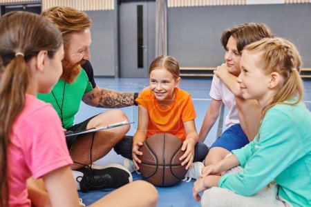 A man teacher with a diverse group of kids sitting around a basketball, engaging in a lively lesson in a bright classroom setting.
