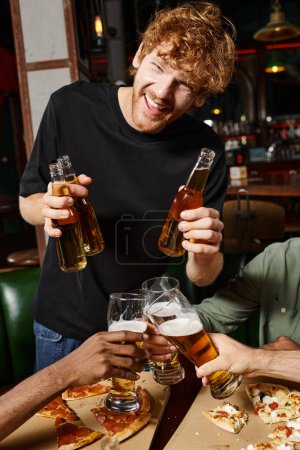 glücklicher rothaariger Mann mit lockigem Haar, der Flaschen mit Bier in der Nähe von Freunden hält, die Gläser in der Bar prosten