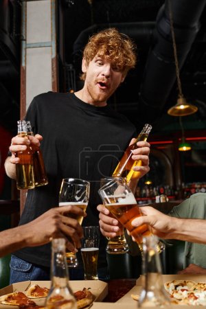 excited redhead man with curly hair holding bottles with beer near friends toasting glasses in bar
