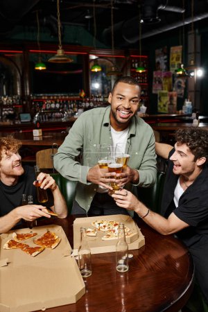 cheerful african american man holding three glasses of beer near male friends and pizza in bar