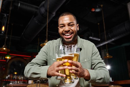glücklicher afrikanisch-amerikanischer Mann mit Hosenträgern, die drei Gläser Bier in der Bar halten und am Wochenende Spaß haben