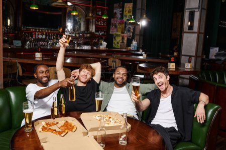 four joyful interracial friends raising bottles and glasses of beer in bar, spending time together