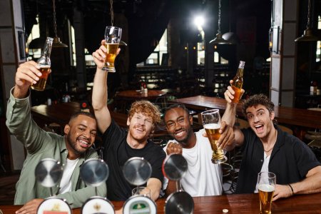 cuatro hombres multiétnicos emocionados levantando vasos de cerveza durante la despedida de soltero, amigos varones en el bar