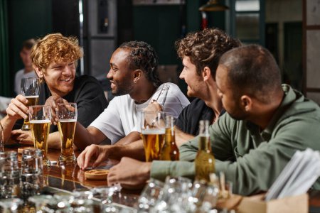 Foto de Hombres multiculturales charlando y bebiendo cerveza, amigos varones felices pasar tiempo juntos en el bar - Imagen libre de derechos