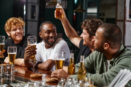 fröhliche multikulturelle Männer, die ihren Freund dabei beobachten, wie er ein Glas Bier anhebt, während er Zeit miteinander in der Bar verbringt