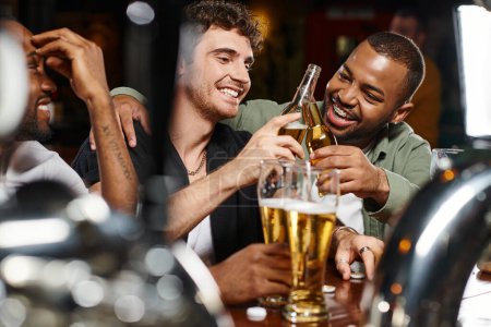 Foto de Tres amigos interracial tostadas con vasos de cerveza mientras pasa tiempo en el bar, amistad masculina - Imagen libre de derechos