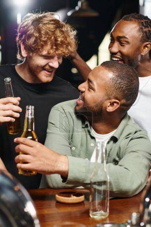 drei fröhliche multiethnische Freunde lächeln und prosten Biergläser zu, während sie Zeit in der Bar verbringen