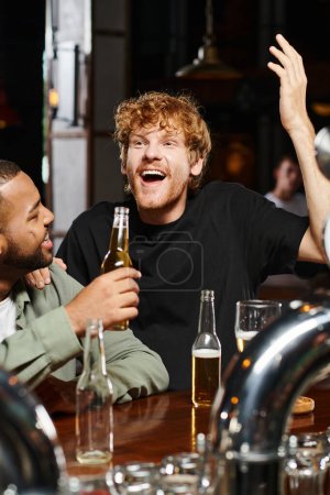 happy redhead man singing near african american friend raising bottle of beer in bar, bachelor party