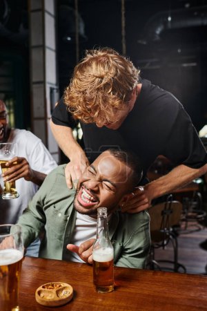 rothaarige Mann kitzelt glücklich afrikanisch-amerikanischen Bräutigam während Junggesellenabschied in Bar, männliche Freundschaft