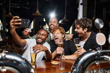 happy multicultural men taking selfie on smartphone while drinking beer in bar, bachelor party