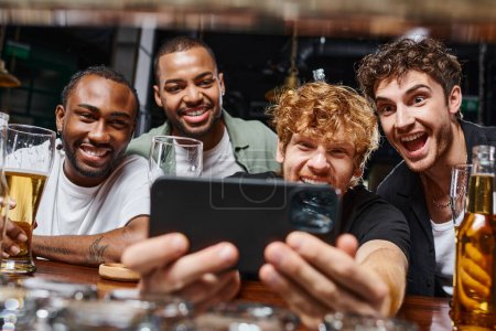 cheerful interracial men taking selfie on smartphone and holding beer during bachelor party in bar