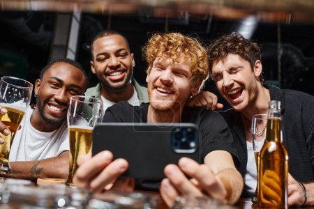 Foto de Hombres interracial positivo tomar selfie en el teléfono inteligente y la celebración de cerveza durante la despedida de soltero en el bar - Imagen libre de derechos