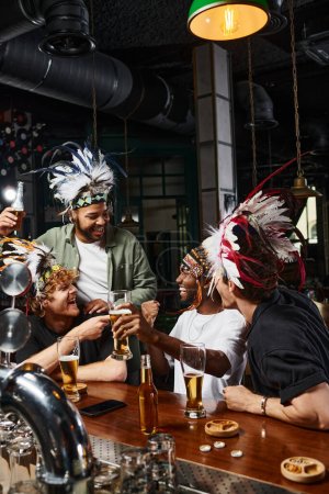 Gruppe aufgeregter männlicher Freunde in Kopfbedeckung mit Federn, die Bier anstoßen und Zeit in der Bar verbringen