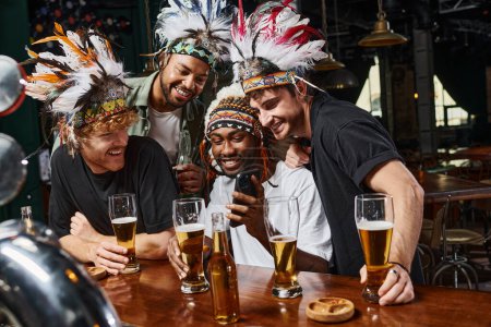 glückliche multikulturelle Männer in Kopfbedeckung mit Federn, die beim Junggesellenabschied in der Bar aufs Smartphone schauen