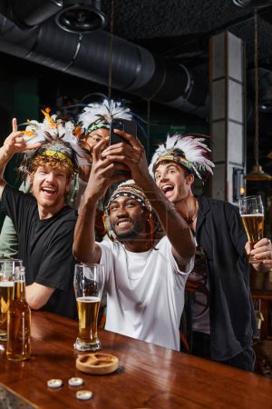 alegres hombres multiculturales en ropa de cabeza con plumas tomando selfie en el teléfono inteligente durante la despedida de soltero