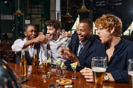 excited multiethnic friends holding tequila shots with lime near glasses of beer on bar counter